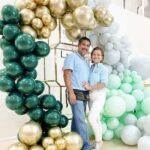 Luz Paz y Omar Bucio junto a un arco orgánico de globos en tonos verdes y dorados.