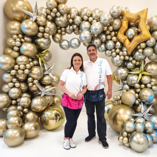 Luz Paz y Omar Bucio posando junto a un maxi arco de globos con técnica orgánica en color dorado y plateado.