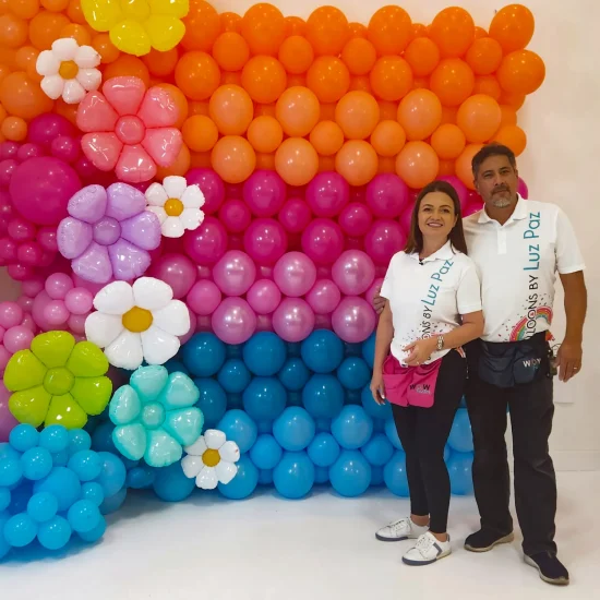 Luz Paz y Omar Bucio junto a pared con globos hecha con técnica Square Pack y flores de globos.